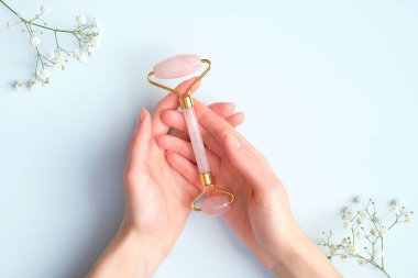 Mavi arka planda gül kuvars masajı yapan kadın el ve çingene çiçekleri. Yeşim taşlı yüz masajı aleti, yaşlanmayı önleyici, kırışıklık önleyici, cilt bakımı aleti.