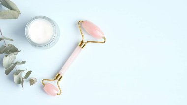 Rose quartz facial massage roller with moisturizer cream and eucalyptus leaf on blue background. Face massager with jade stone, anti-aging, anti-wrinkle beauty skincare tool clipart
