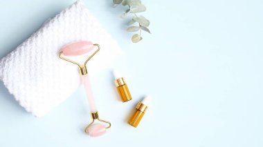 Yüz için Jade silindiri ve mavi arka planda havlusu olan gerekli yağ. Düz konum, üst görünüm, kopyalama alanı. Gül kuvars yüz masajı aracı, cilt bakımı için güzellik aracı