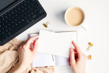 Ekose, kahve ve dizüstü bilgisayarla ofis masasının üzerinde boş kâğıt maketi tutan kadın elleri. Hygge, rahat bir ortam, rahat bir konsept. Düz yatış, üst görünüm