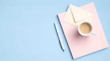 Pembe not defteri, bir fincan kahve, zarf, mavi arka planda kalem olan en az çalışma alanı. Modern ofis masası, iş veya eğitim konsepti