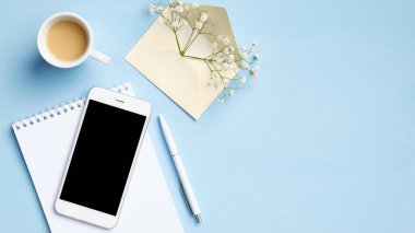 Bir fincan kahveli kadınsı çalışma alanı, boş ekran maketli akıllı telefon, not defteri, çiçekli zarf, mavi arka planlı kalem. Merkez ofis masası, düz yatak, üst manzara.