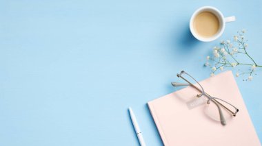 Pembe defterli modern ofis masası, kalem, bardak, kahve, mavi arka planda çiçekler. En az kadınsı çalışma alanı, iş ve eğitim konsepti. Düz yatış, üst görünüm