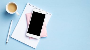 Akıllı telefonlu, boş ekranlı modern ofis masası, bir fincan kahve, boş kağıt not defteri, mavi arka planda belge. Kopyalama alanı olan üst görünüm, düz yatay