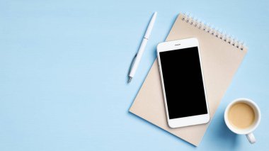 Kağıt defter, kalem, siyah ekranlı akıllı telefon, mavi arka planda bir fincan kahve. Fotokopi alanı olan üst görünüm, düz. Asgari biçimdeki kadınsı çalışma alanı