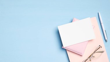 Düz uzanış, üst görünüm, pembe defterli, gözlüklü, belgeli, mavi arka planda kalem ve boş kağıt kartlı kadınsı çalışma alanı. Modern ofis masa konsepti