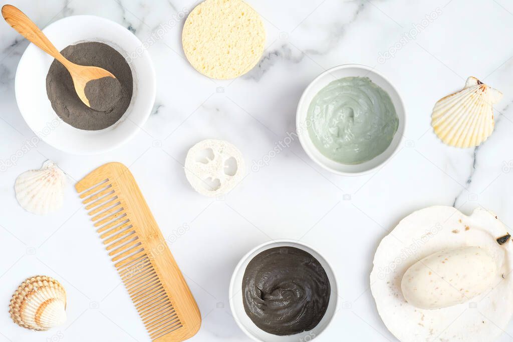 Face clay masks in bowls with hair comb, loofah sponge, homemade soap and seashells on marble background. Flat lay, top view. SPA natural organic cosmetics for face treatment, skin care