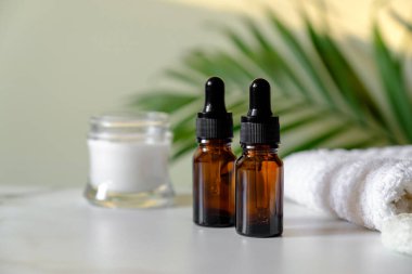 Tuvaletteki mermer masada iki tane temiz damlatıcı şişesi, vücut kremi ve havlu. Arka planda palmiye yaprağı var. Temel yağ veya serum için kahverengi kehribar cam kaplar. SPA doğal organik kozmetik seti.