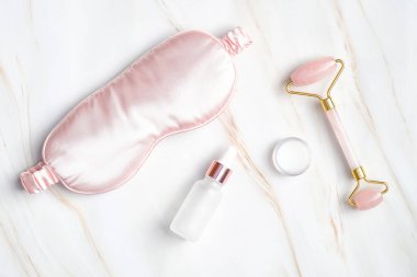 Uyuyan göz maskesi, serum düşürücü şişesi, nemlendirici krem mermer arka planda yüz masajı silindiri. Gece yarısı cildi rutin konsepti