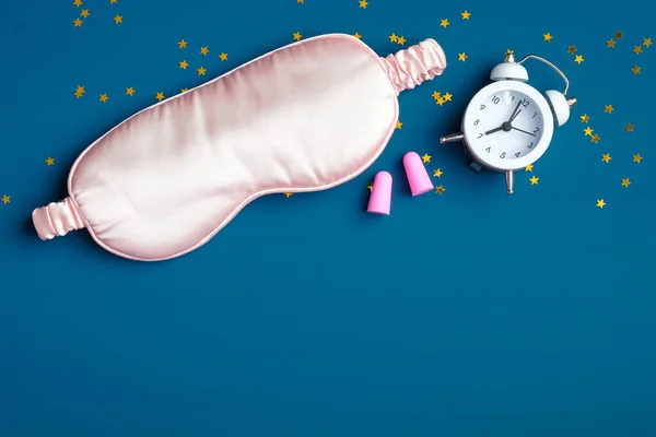 Healthy sleeping concept. Sleep eye mask with pink ear plugs and alarm clock on dark blue background. Flat lay, top view.