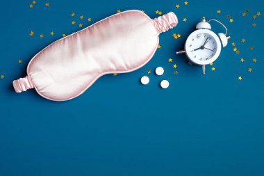 Pembe göz maskesi, çalar saat, koyu mavi arka planda uyku ilacı. Düz yerde, üst tarafta. İyi geceler, uyku hijyeni, uykusuzluk tedavisi konsepti..