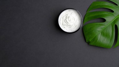 Kozmetik kremalı kavanoz ve siyah arka planda tropikal canavar yaprağı. Vücut, el ve yüz bakımı için doğal organik güzellik ürünü. Düz yerde, üst tarafta. Cilt bakımı kavramı.