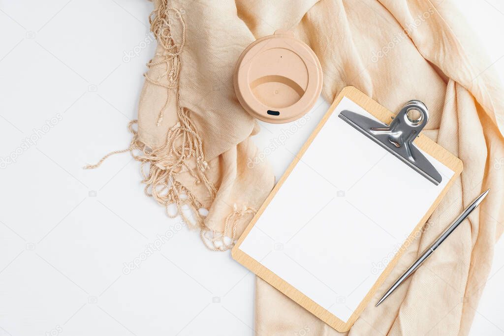 Cozy home office desk with beige blanket, cup of coffee, clipboard with empty paper note on white background. Flat lay, top view. Hygge or nordic style feminine workspace.