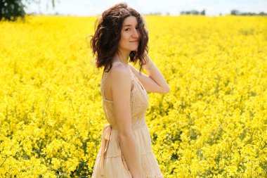 Yaz günü sarı tarlada kıvırcık saçlı neşeli beyaz kız. Bej elbiseli mutlu genç kadın doğanın tadını çıkarıyor, kameraya bakıyor. Yakın plan portre.