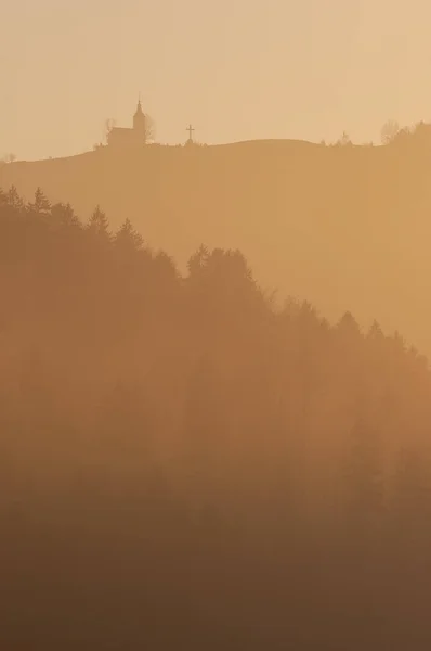 Piękny widok na stoków leśnych i kościoła na szczycie wzgórza w wieczornej mgle — Zdjęcie stockowe