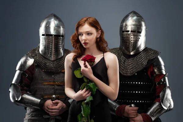 Siyah elbiseli güzel bir kız ve elinde kırmızı bir gül zırhlı iki şövalyenin yanında.
