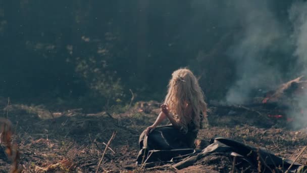 Молодая женщина сидит на земле возле горящего леса — стоковое видео