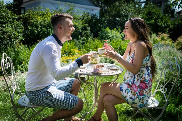 Jovem casal feliz senta-se à mesa e segura copos com chá no jardim — Fotografia de Stock
