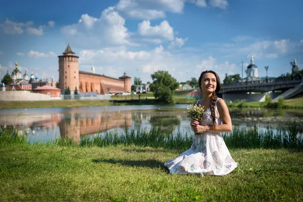 Jovem mulher com flores em vestido leve senta-se na grama — Fotografia de Stock