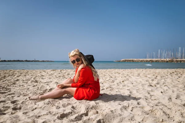 Joven mujer atractiva en vestido rojo brillante se sienta en la playa — Foto de Stock