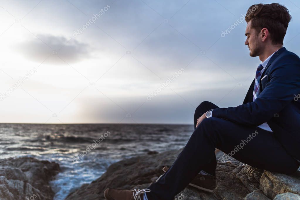 Pensive man in suit sits on the rocks near sea and thinking. Young guy