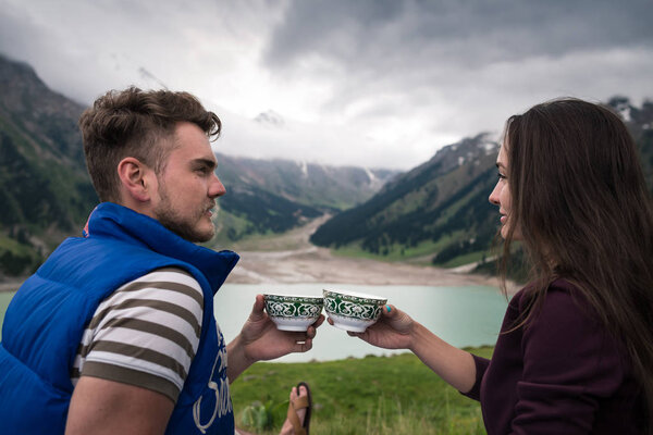 Young modern couple sits near beautiful lake and holds in hands traditional cups
