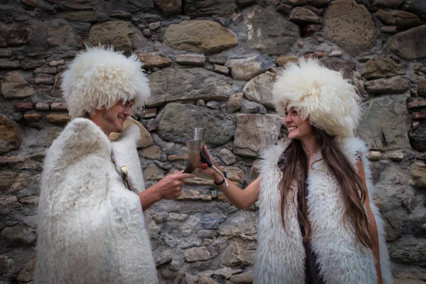 Funny couple in national woolen clothes are drinking from traditional glasses — Stock Photo, Image