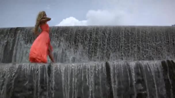 大きな滝の上に行くオレンジのドレスで巻きブロンドの髪の若い美しい女性 緑の森 ヤシと背景に曇り空 — ストック動画