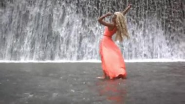 Büyük şelalenin arka planında poz veren güzel bir kadın. Turuncu elbiseli kız el kaldırıyor, kameraya bakıyor ve anın tadını çıkarıyor. Arkadan görüntüle.