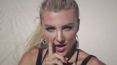 Sarı saçlı, kısa siyah elbiseli ve mayolu gizemli genç bir kadın beyaz kumsalda tek başına oturuyor ve poz veriyor..