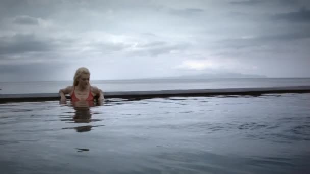 严肃的年轻女子 金发碧眼 身穿红色游泳衣 悠闲自在地在努沙佩尼达岛上无边无际的游泳池里 背景上的海洋 天空和天空 — 图库视频影像