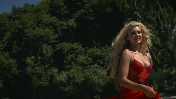 年轻而积极的女人 金发碧眼 穿着优雅飘逸的红色长裙 走在沙滩上 面带微笑 背景上的绿树和棕榈树 — 图库视频影像