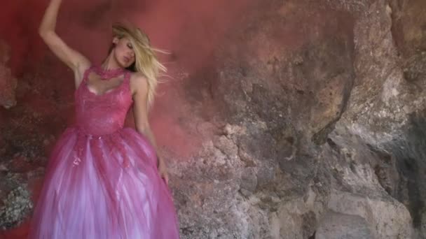 若いです魅力的な女性とともにブロンド髪でピンクふわふわドレス上の砂の上に立っていると大きな岩の背景に厚い煙でポーズ — ストック動画