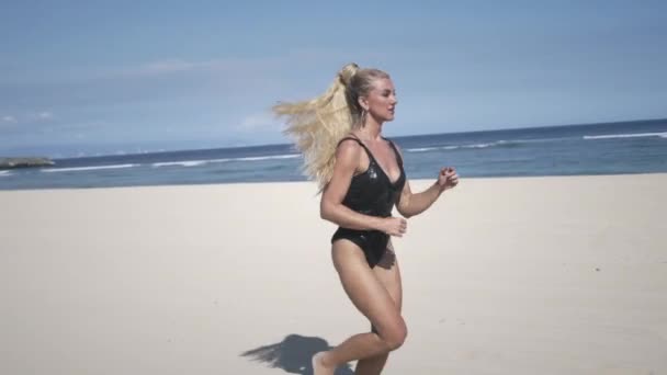 年轻女子 金发碧眼 身穿黑色的小礼服 身穿泳衣 在海边的白沙滩上奔跑 纵观海岸线和天空 — 图库视频影像