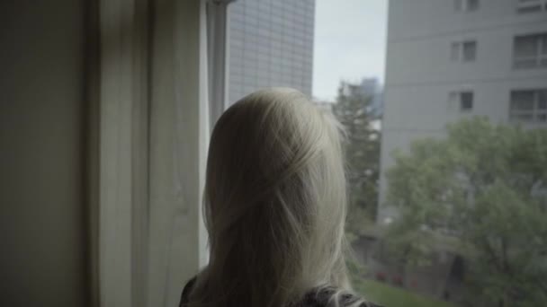 建物の眺めと窓の近くにポーズブロンドの髪を持つ若い眠い女性 スタイリッシュなアパートで夜更かしをする黒人の少女 バックビュー — ストック動画