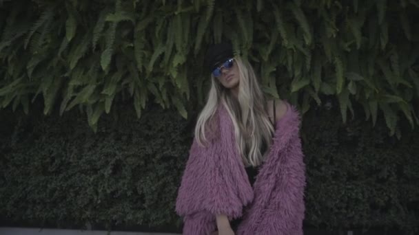 年轻而奇怪的女人 金发碧眼 身穿粉色皮大衣 戴着太阳镜 站在绿叶和植物丛生的背景上 — 图库视频影像