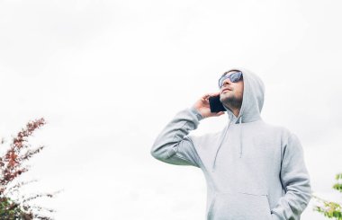 Gri eşofman altı ve mavi güneş gözlüklü genç adam ellerinde cep telefonu ve beyaz bir arka planda gülümsüyor.