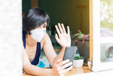 Kadın, evindeki pencereden video görüşmesi yapıyor ve koronavirüs karantinası sırasında maske takıyor.
