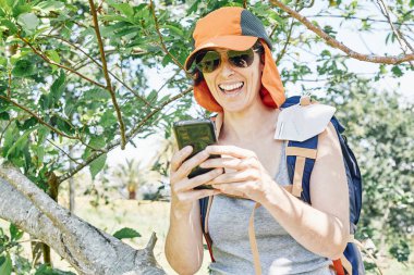 Sırt çantası ve maskesiyle yürüyüş gününden sonra cep telefonuyla sohbet ederken gülen bir kız.