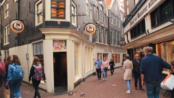 AMSTERDAM, Netherlands - October 2019: Local and tourists strolling through the narrow little streets of the city. In the foreground a storefront of a cheese sale shop — Stock Video