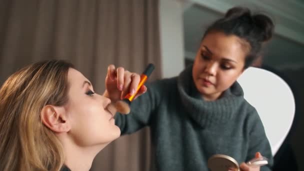 Processus de maquillage. Maquilleuse travaillant au pinceau sur le visage du mannequin. Portrait de jeune femme blonde dans le salon de beauté intérieur. Appliquer ton sur ton sur la peau — Video