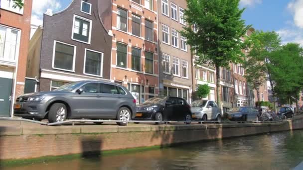 Amsterdam, Netherlands - September 2019: cyclist rides along the narrow street of the capital. Parked cars stand near the canal — Stock Video