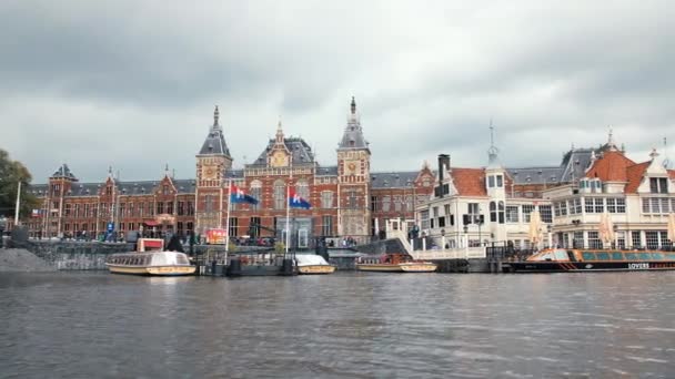 Amsterdam, Netherlands - September 2019: Central Station, Canal view of the cityscape. Кілька прапорів столиці махають вітром. Пришвартований великий туристичний круїзний човен. — стокове відео