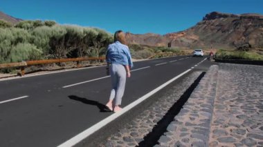 Kot ceketli ve çizgili pantolonlu kız dağlık arazide asfalt bir yol boyunca gidiyor..