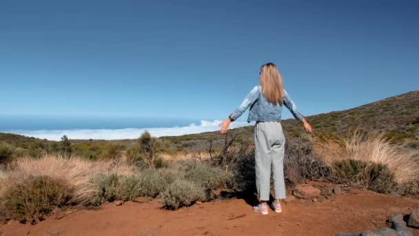 Blond kvinna i jeansjacka och randiga byxor står på ett högt berg ovanför molnen och lyfter upp händerna. Begreppet frihet, avkoppling, inspiration — Stockvideo