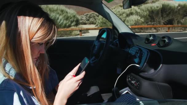 Vonzó szőke nő farmerdzsekiben okostelefont használ, miközben egy autó anyósülésén ül. A napsugarak megvilágítják a modell haját. — Stock videók