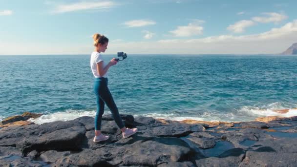 Femme blonde blogueur influenceur en jeans et blanc T-shirt tourne la vidéo sur smartphone en utilisant stabilisateur de cardan. Sur la rive volcanique de l'océan. Concept de création de contenu pour les réseaux sociaux, cinématographie . — Video