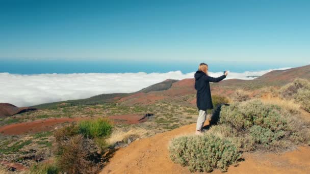 Çekici sarışın kadın akıllı telefon kullanarak selfie çekiyor. Vadinin yukarısında bulut denizi ve dağ sırtlarını kaplayan orman ile güzel bir manzara. Etkilendirici kavramı sosyal ağlar için içerik oluşturur. — Stok video