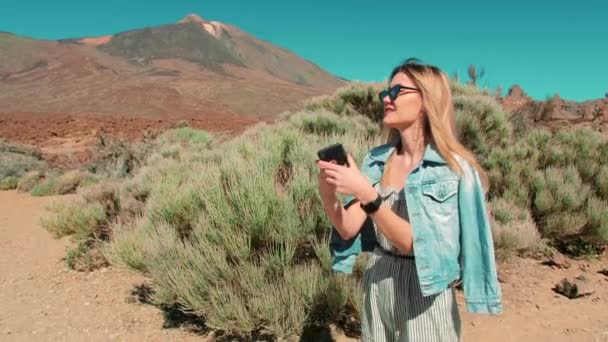 Kot ceketli ve güneş gözlüklü bir kadın cep telefonu kullanıyor, doğanın fotoğraflarını çekiyor. Volkanik dağ ve kayaların arkasındaki çöl. Etkilenen kişi kavramı sosyal ağlar için içerik üretir — Stok video