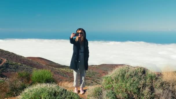 Ελκυστική ξανθιά γυναίκα κάνει selfie χρησιμοποιώντας ένα smartphone. Όμορφο τοπίο με θάλασσα από σύννεφα πάνω από την κοιλάδα και δάσος που καλύπτει βουνοκορφές. έννοια της έμπνευσης, της ελευθερίας — Αρχείο Βίντεο